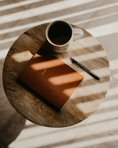 Leather journal and coffee mug on a round wooden table with dappled morning light