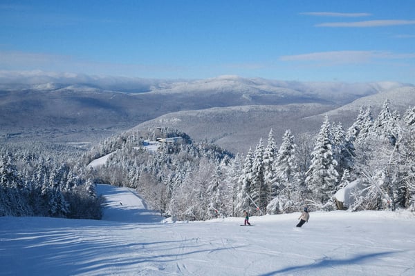 Two skiers carving fresh powder at Loon Mountain with snow-covered pines and blue sky