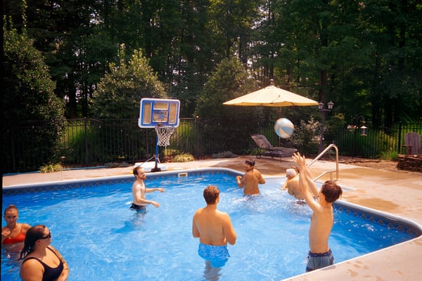 Group of friends playing pool volleyball in a backyard pool surrounded by trees on a summer day