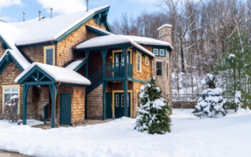Brick and wood cottage with blue trim and snow-covered roof surrounded by snow and bare trees
