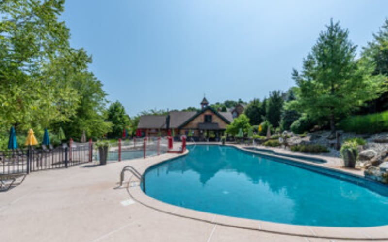 Curved swimming pool with blue water, concrete deck, metal fence, and house with trees in background