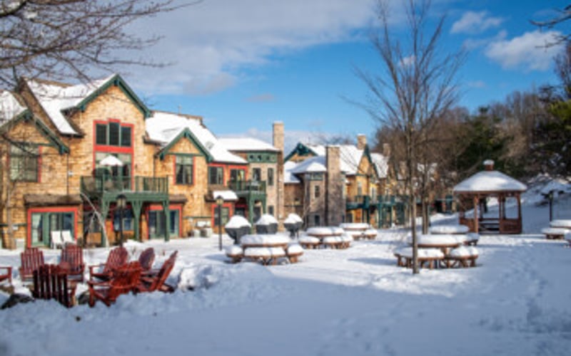 Snow-covered town square with yellow stone buildings, bare trees, and gazebo on sunny winter day