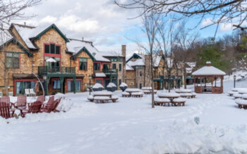 Snow-covered village square with stone buildings, bare trees, and wooden benches