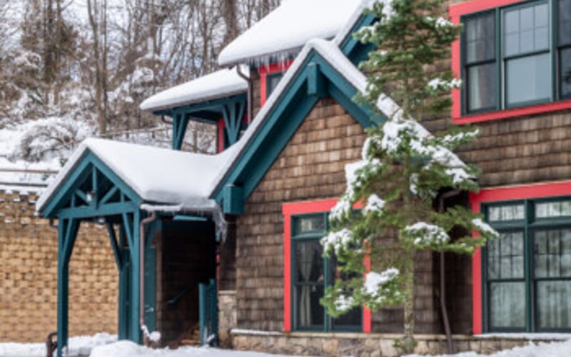 Snowy cottage with blue-trimmed entrance and red-trimmed windows, surrounded by bare trees