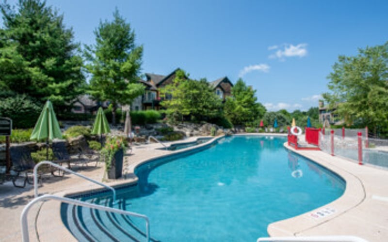 Resort swimming pool with curved shape, lounge chairs, umbrellas, and residential buildings surrounded by trees