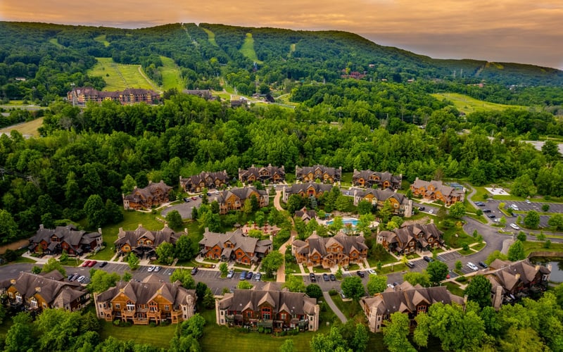 Aerial view of residential neighborhood with brick homes nestled in forested valley beneath ski slopes at sunset