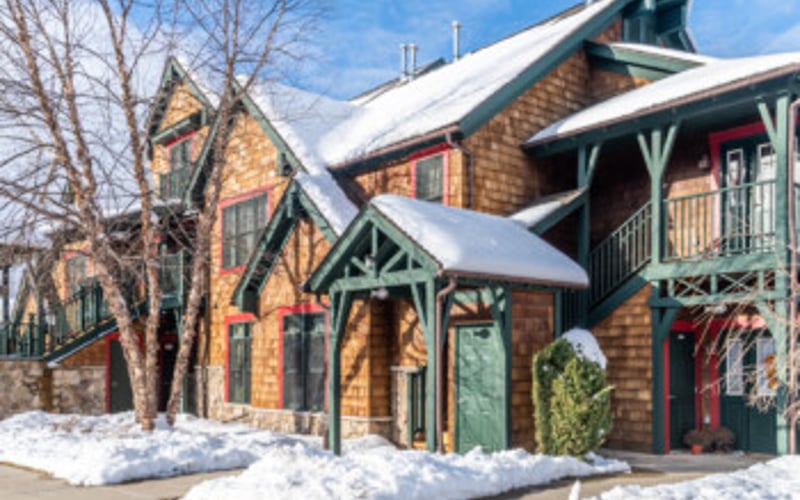 Brick cottage with snow-covered peaked roof and green trim, bare trees nearby