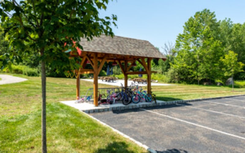 Wooden picnic shelter with metal roof in grassy park area next to paved parking lot