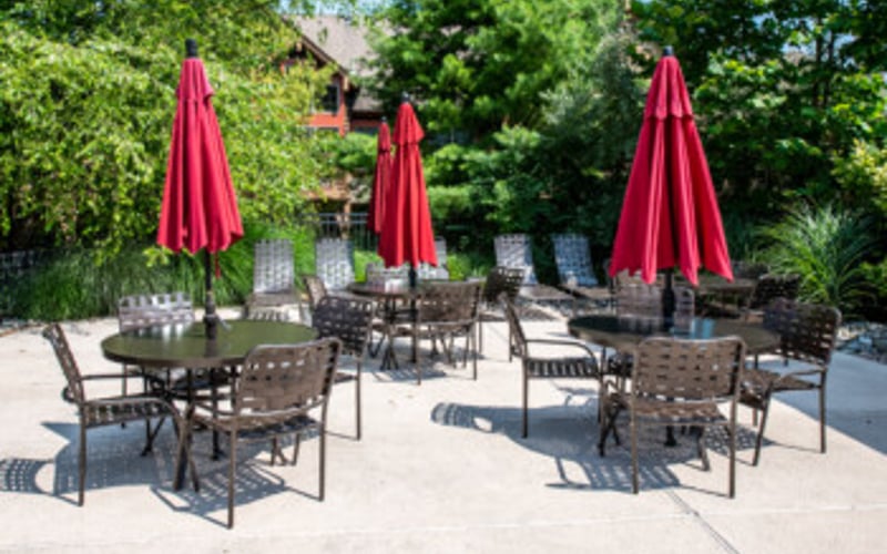 Outdoor patio with multiple dining tables, chairs, and red umbrellas surrounded by green trees