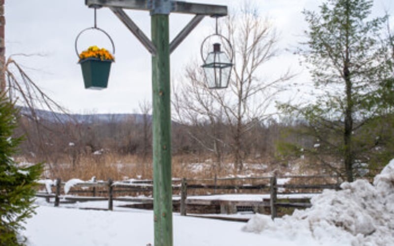 Green wooden post with hanging flower baskets overlooking snowy landscape and bare trees